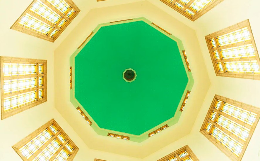 Inside the Bahá'í Temple in Kampala, featuring a pale blue dome, wooden pews, and soft light filtering through multi-colored glass windows, creating a serene and meditative atmosphere."