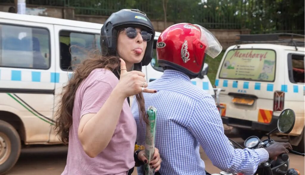 A white woman riding on a boda boda through a bustling Ugandan street, surrounded by local shops and traffic, capturing the adventurous spirit of travel. This image represents the experience of exploring Uganda at different times of the year. Best times to visit Uganda depend on the season and planned activities.